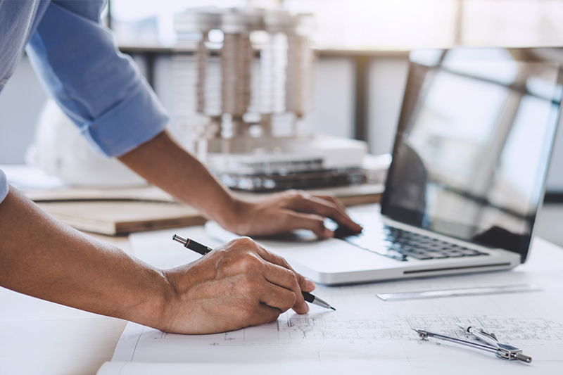 Person reviewing architectural plans while working on a laptop at a drafting table.